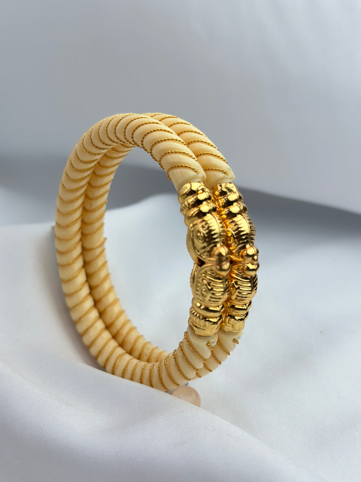 A double-strand braided bracelet made of cream-colored cord with a prominent textured gold clasp, displayed on a soft white fabric background.