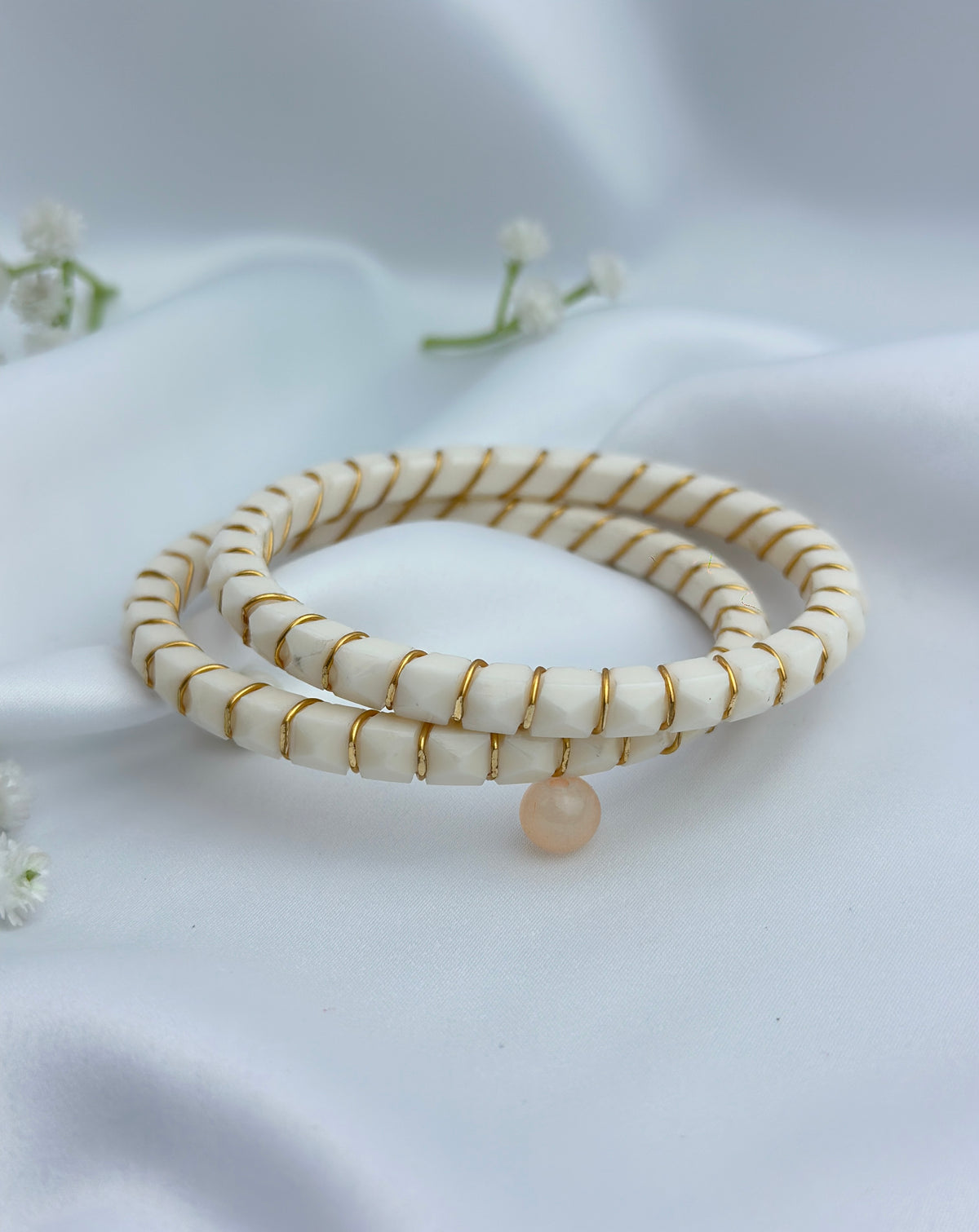 A pair of double-strand beaded bracelets with cream-colored beads and thin gold wire wrapping, featuring a small peach-colored pearl charm, displayed on a white satin fabric background.