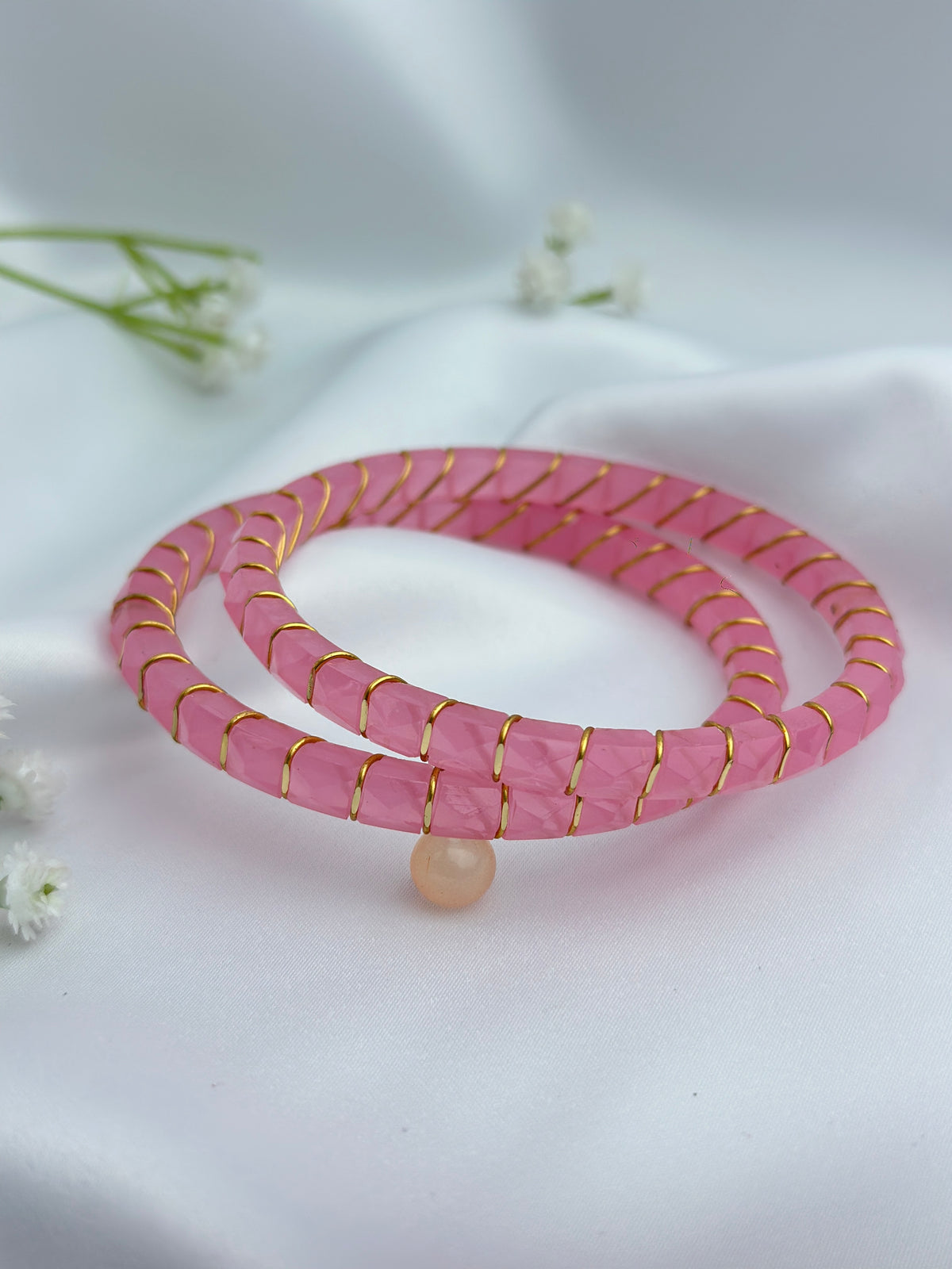 A pink beaded wrap bracelet with gold wire wrapping and a small pearl bead, displayed on a white satin fabric background with small white flowers.