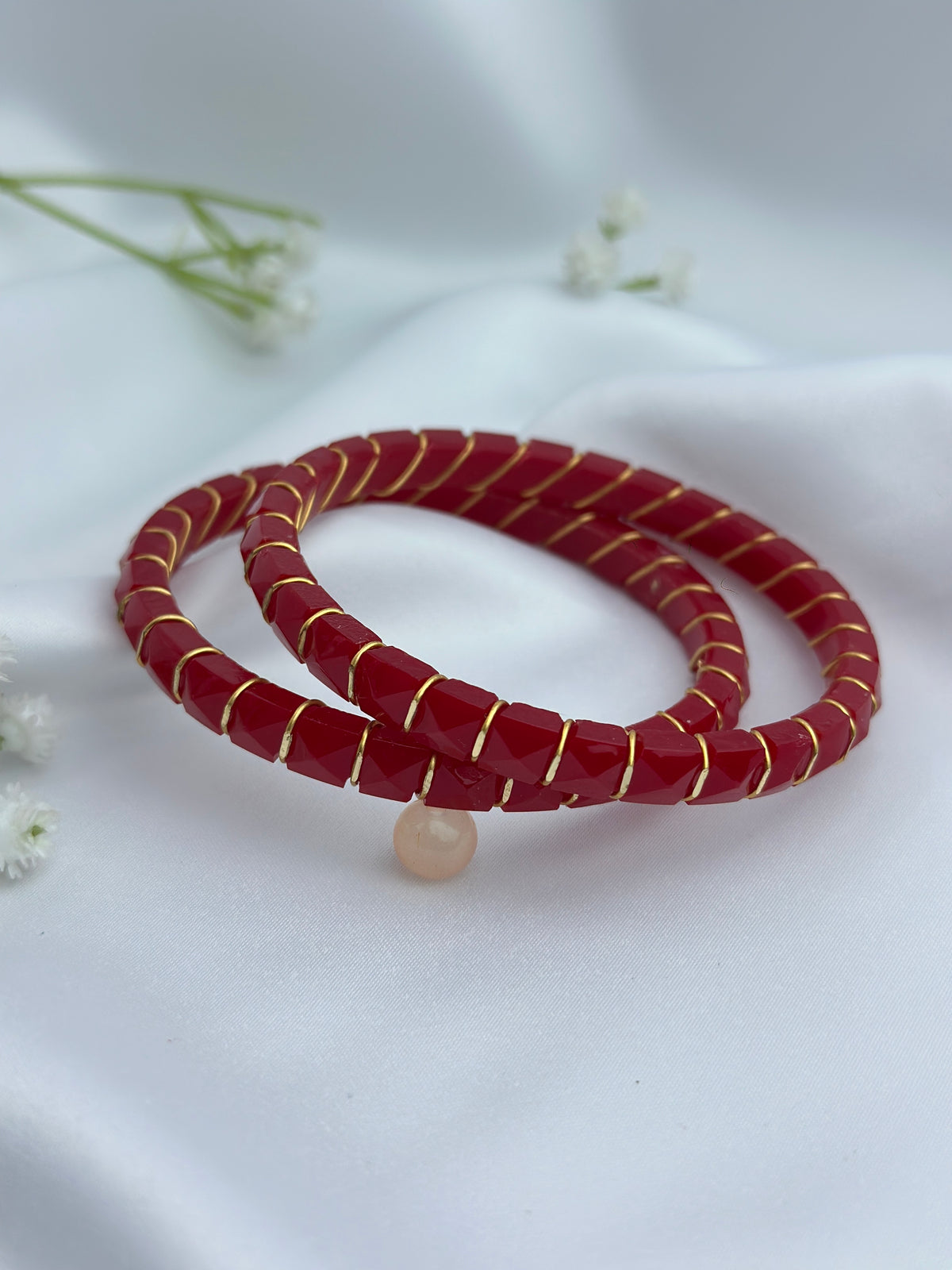 Two red beaded bracelets with gold twisted detailing and a small peach bead charm, displayed on a white fabric background with small white flowers.