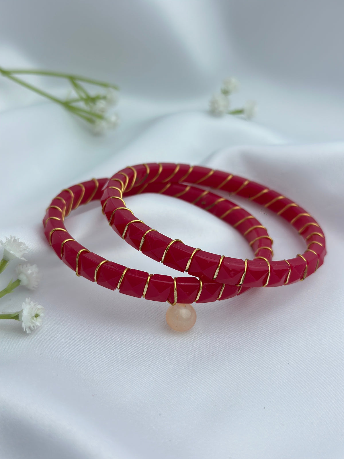 Two red beaded bracelets with gold wire wrapping and a small peach stone charm, displayed on a white fabric background with small white flowers.