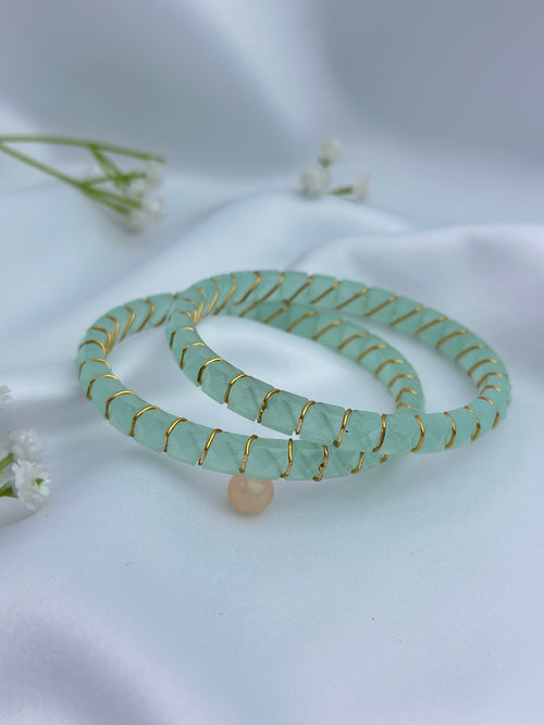 Two light green jadeite beaded bracelets with gold wire wrapping and a small peach bead charm, displayed on a white satin fabric background with small white flowers.