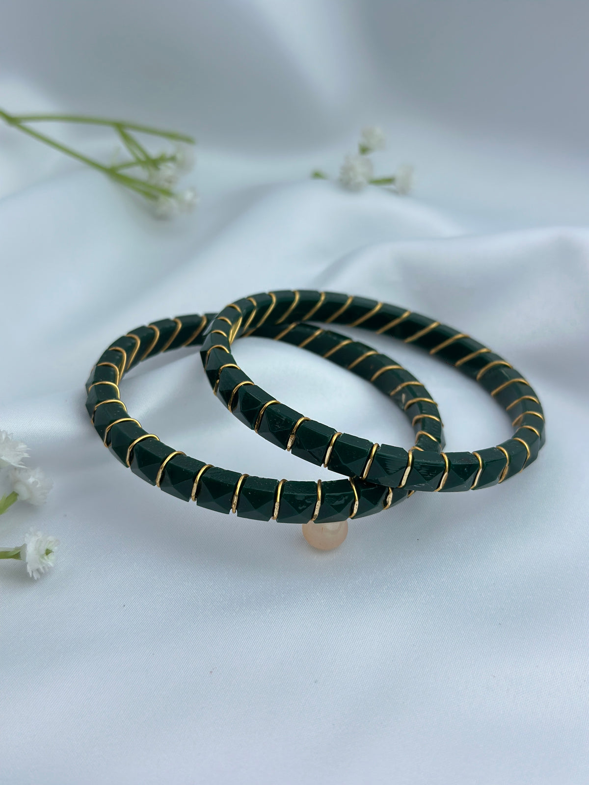 Two dark green beaded bracelets with thin gold wire wrapped accents, displayed on a soft white fabric background with small white flowers.