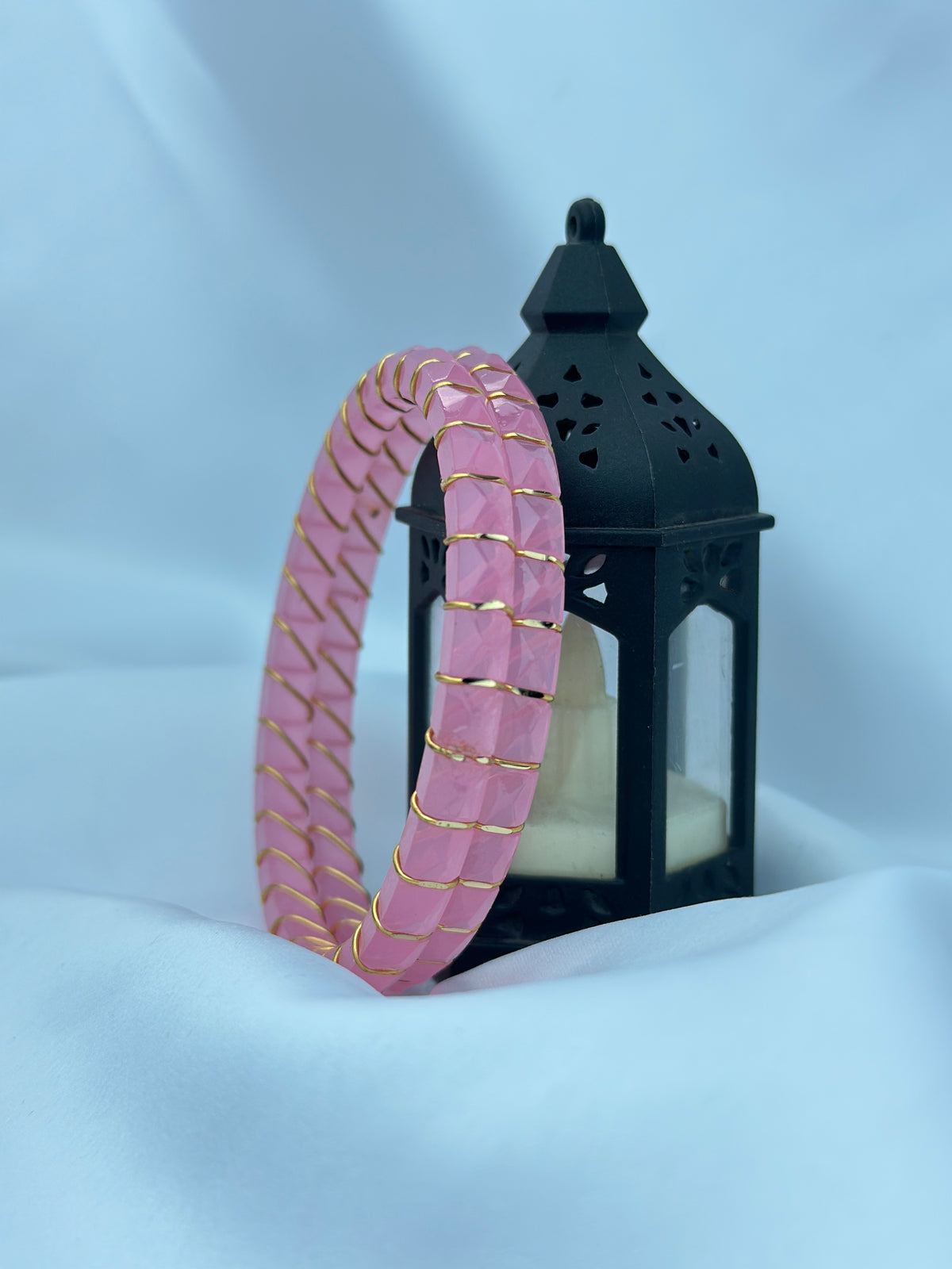A pink segmented bangle bracelet with gold trim, draped over a black decorative lantern on a light blue fabric background.