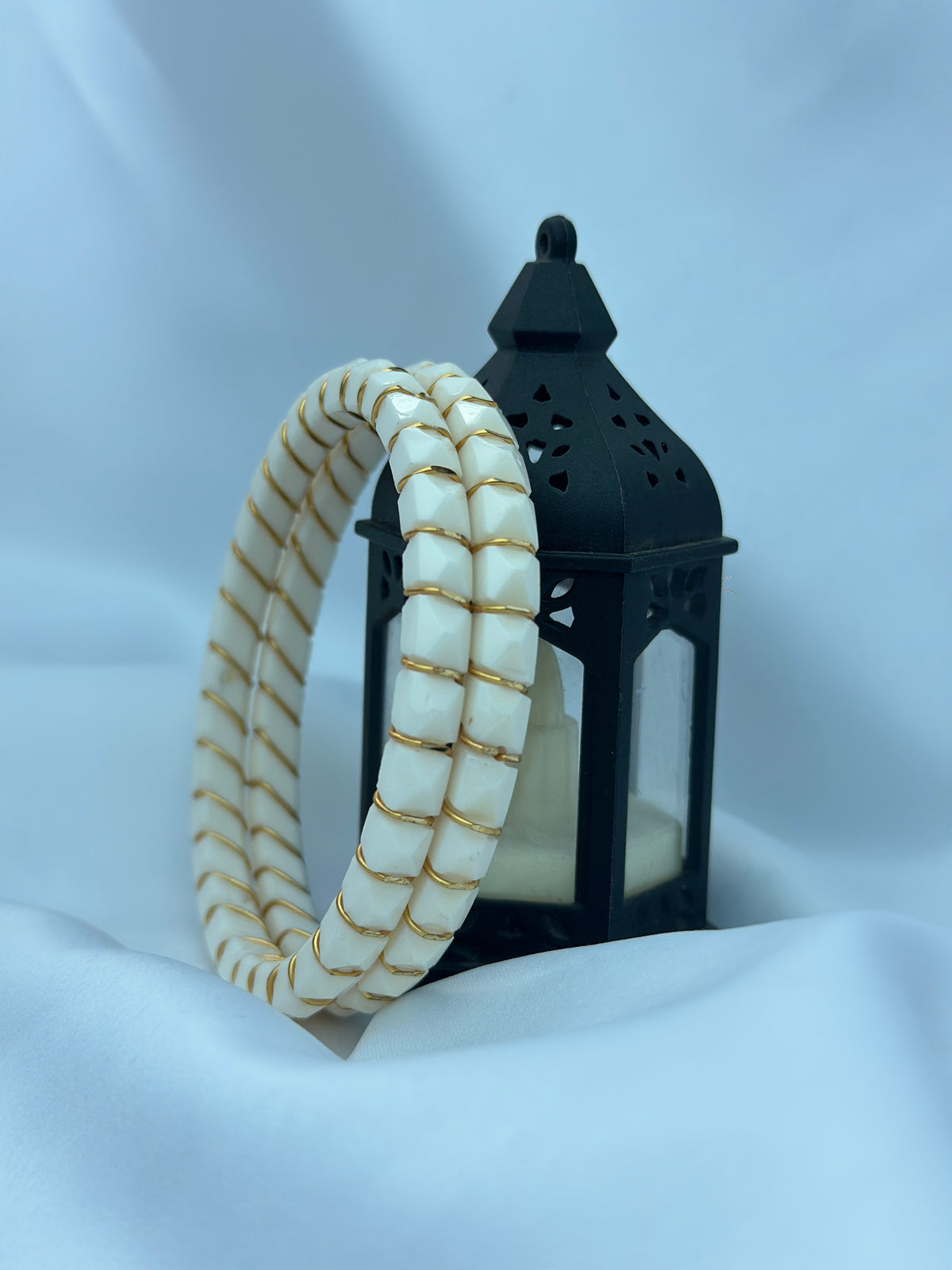 Two bangles displayed in front of a black lantern candle holder on draped white fabric. One bangle is cream-colored with gold stripes, and the other is cream with white rectangular segments separated by thin gold lines.