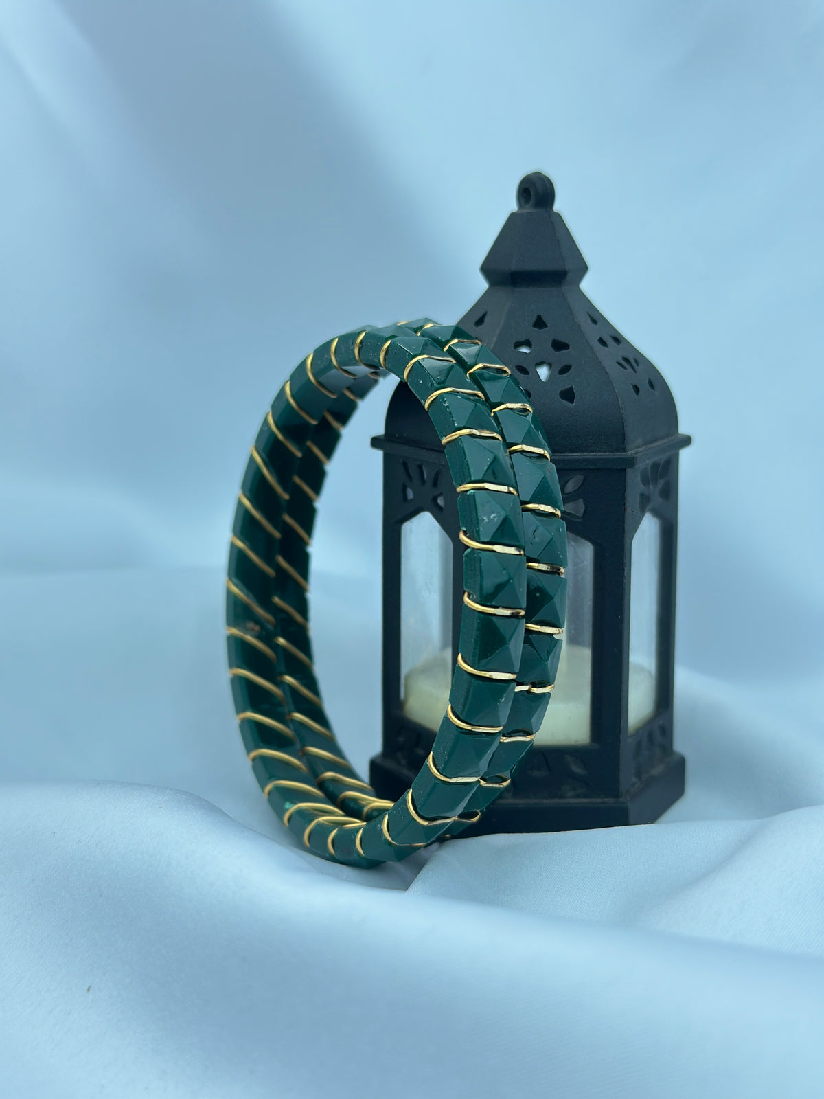A pair of dark green beaded bracelets with gold trim, displayed in front of a black decorative lantern on a light blue fabric background.