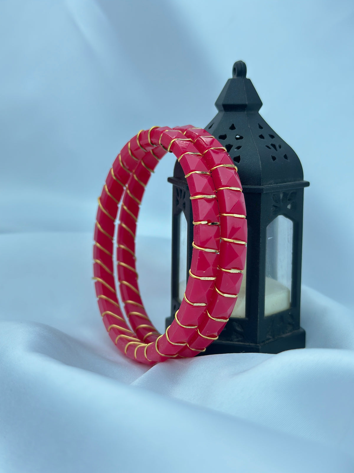 Two red beaded bangles with gold trim, displayed in front of a black decorative lantern on a draped light blue fabric background.