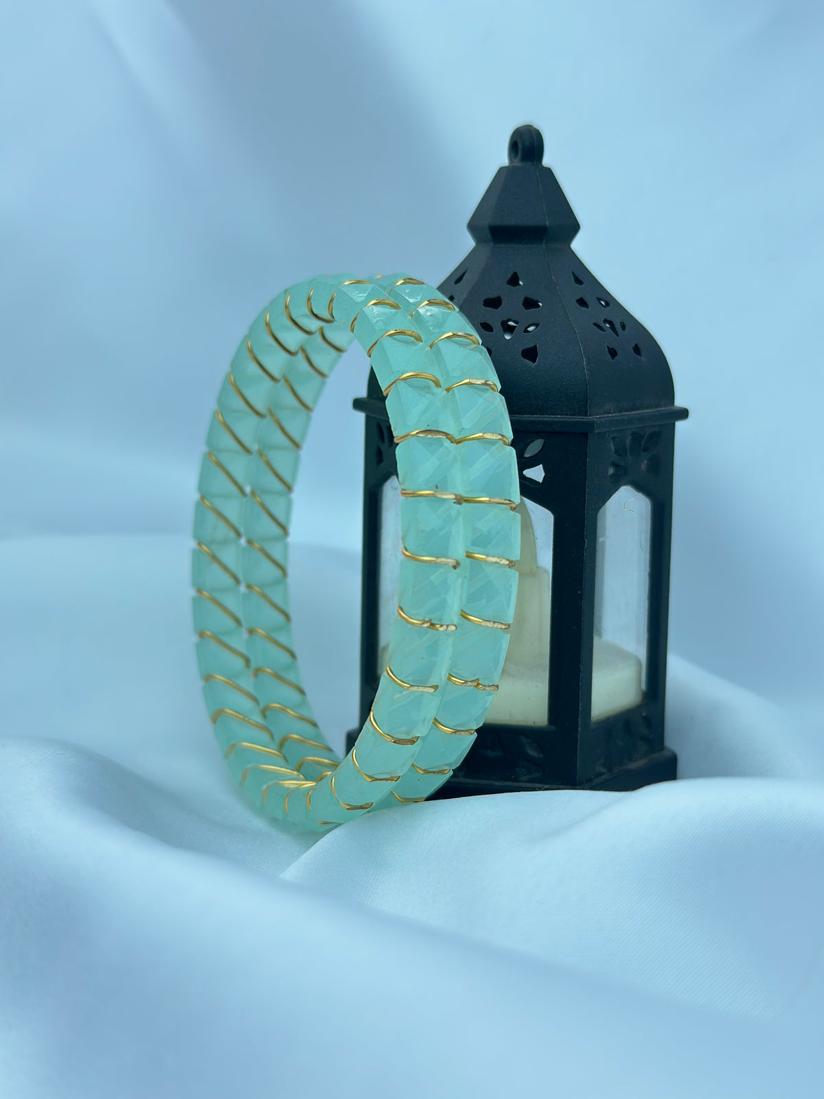 A mint green stone bangle bracelet with gold wire wrapping, displayed next to a black decorative lantern on a draped light blue fabric background.