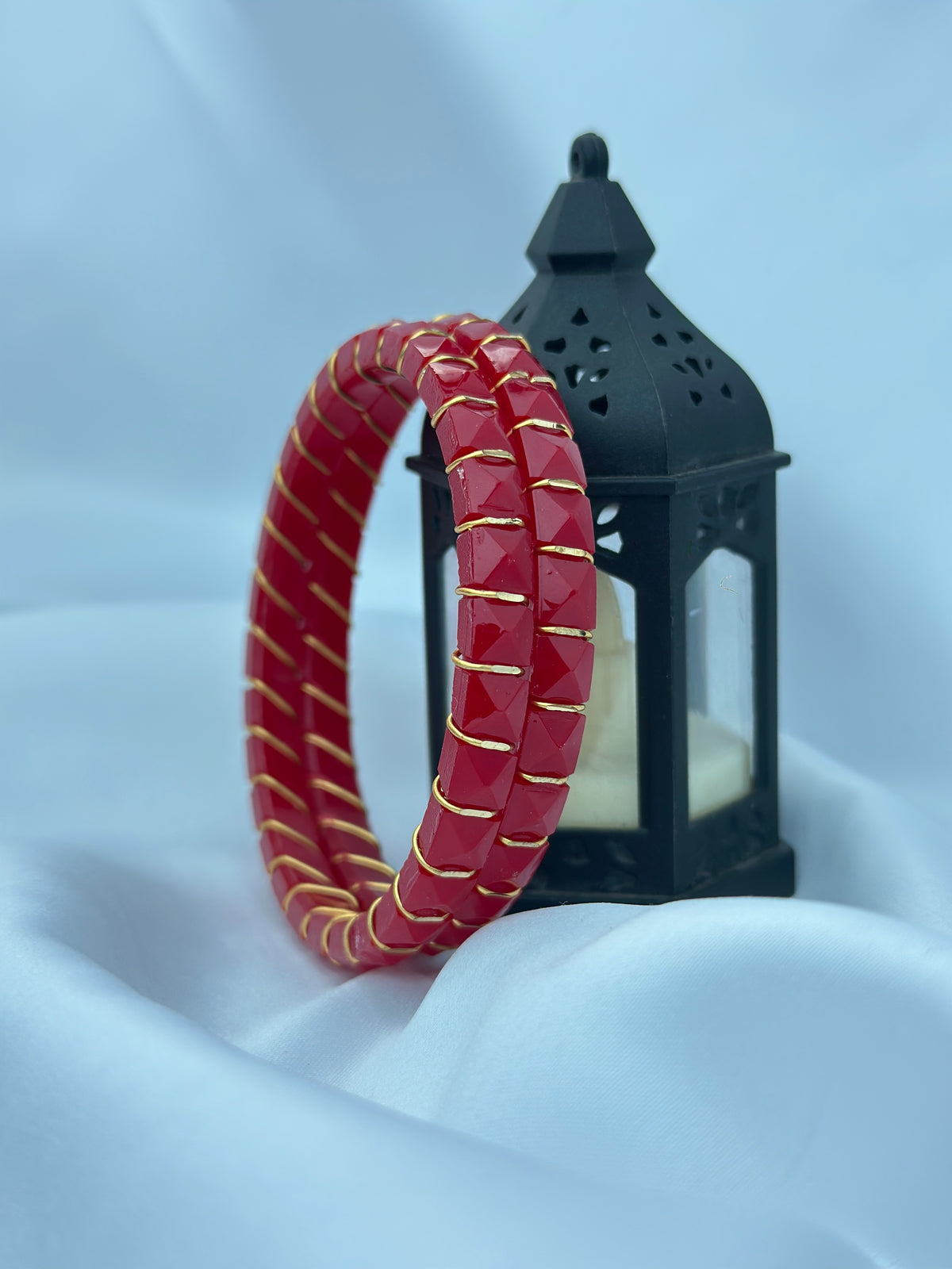 A red beaded bracelet with gold trim is displayed in front of a black decorative lantern on a soft white fabric background.
