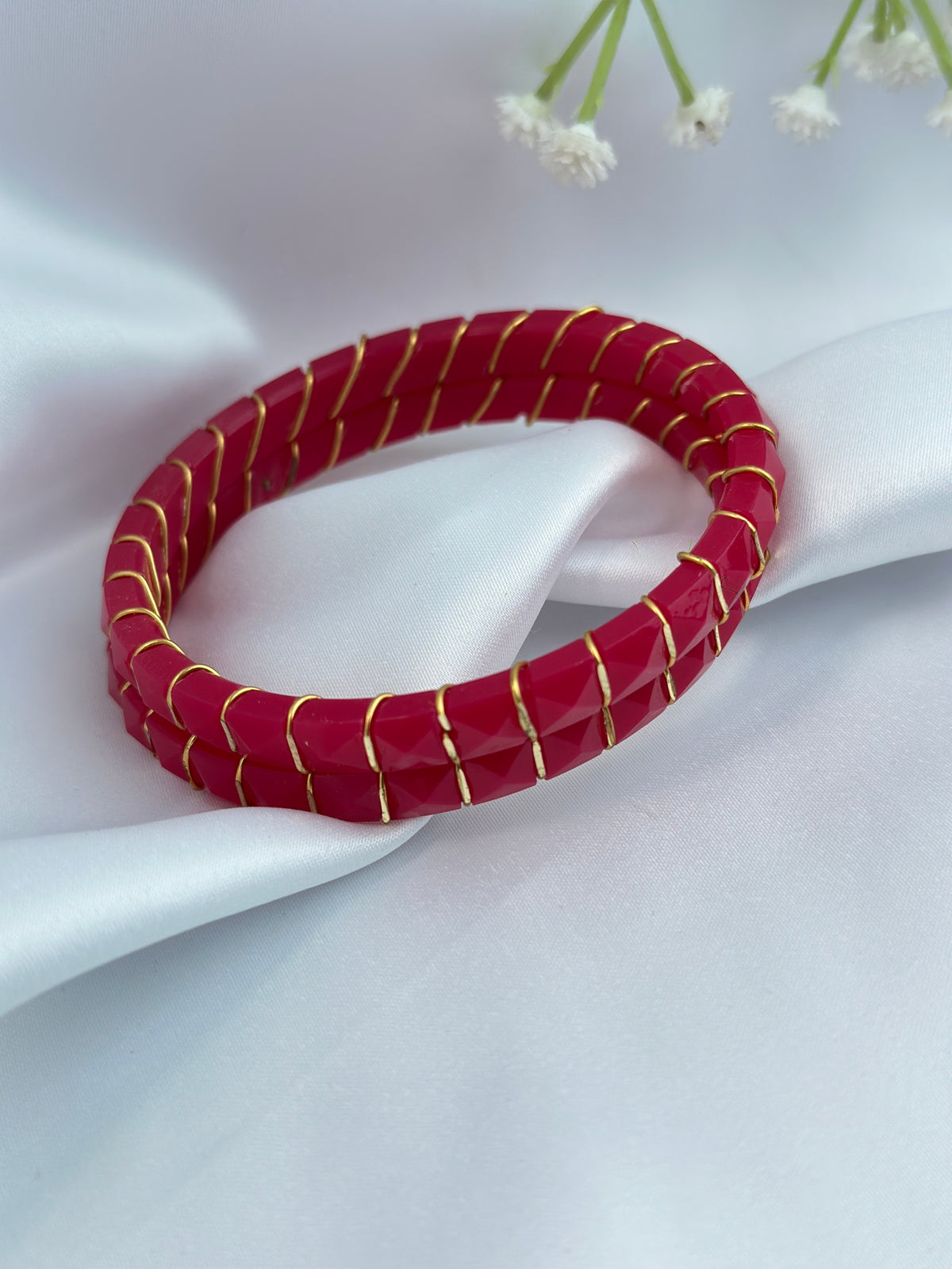 A red beaded bangle bracelet with thin gold metallic accents, displayed on draped white satin fabric with small white flowers in the background.