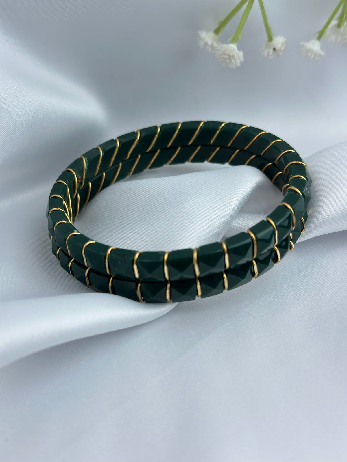 A dark green beaded stretch bracelet with thin gold wire wrapped around it, displayed on a draped white satin fabric background with small white flowers in the corner.