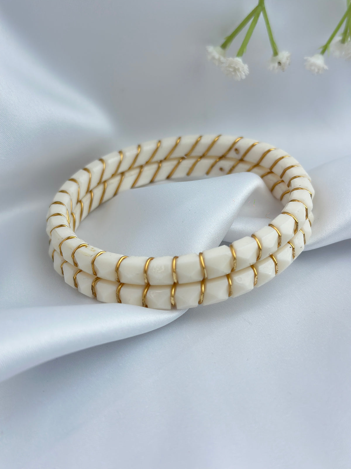 A cream-colored beaded bracelet with gold wire wrapped details, displayed on a draped white satin fabric background with small white flowers in the corner.