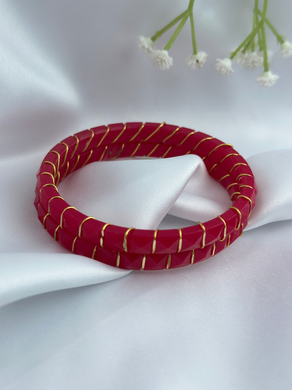 A red beaded bangle bracelet with thin gold metallic stripes, displayed on a white satin fabric background with small white flowers in the upper corner.