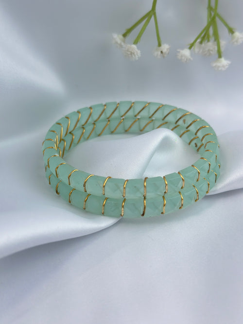 A light green beaded stretch bracelet with gold wire wrapping, displayed on a draped white satin fabric with small white flowers in the background.