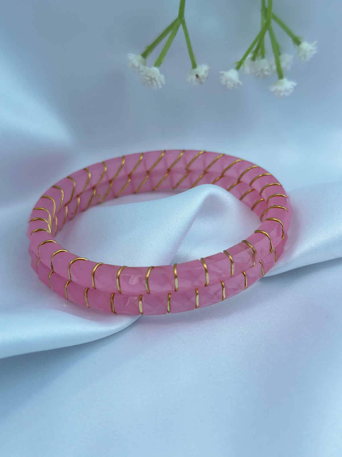 A pink stone bangle bracelet with gold wire wrapping, displayed on a draped white satin fabric with small white flowers in the background.