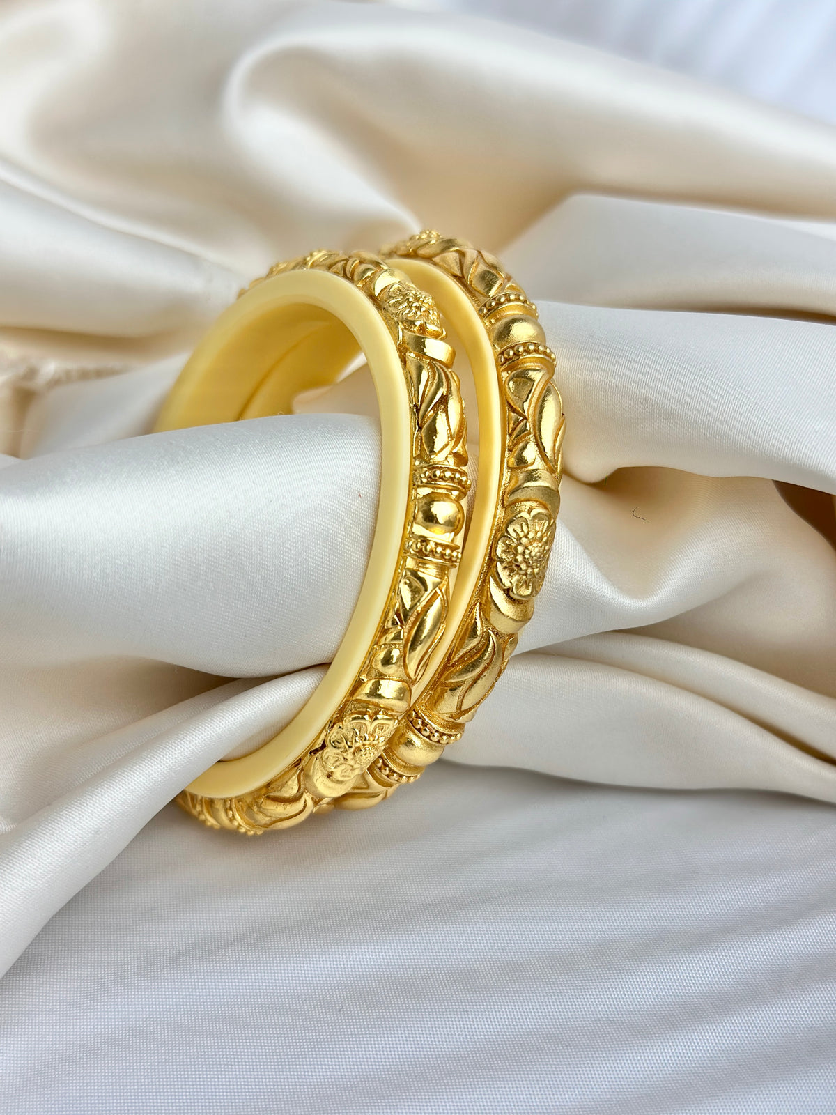 A pair of gold-toned traditional rings with intricate floral and geometric engravings, displayed on draped cream satin fabric.