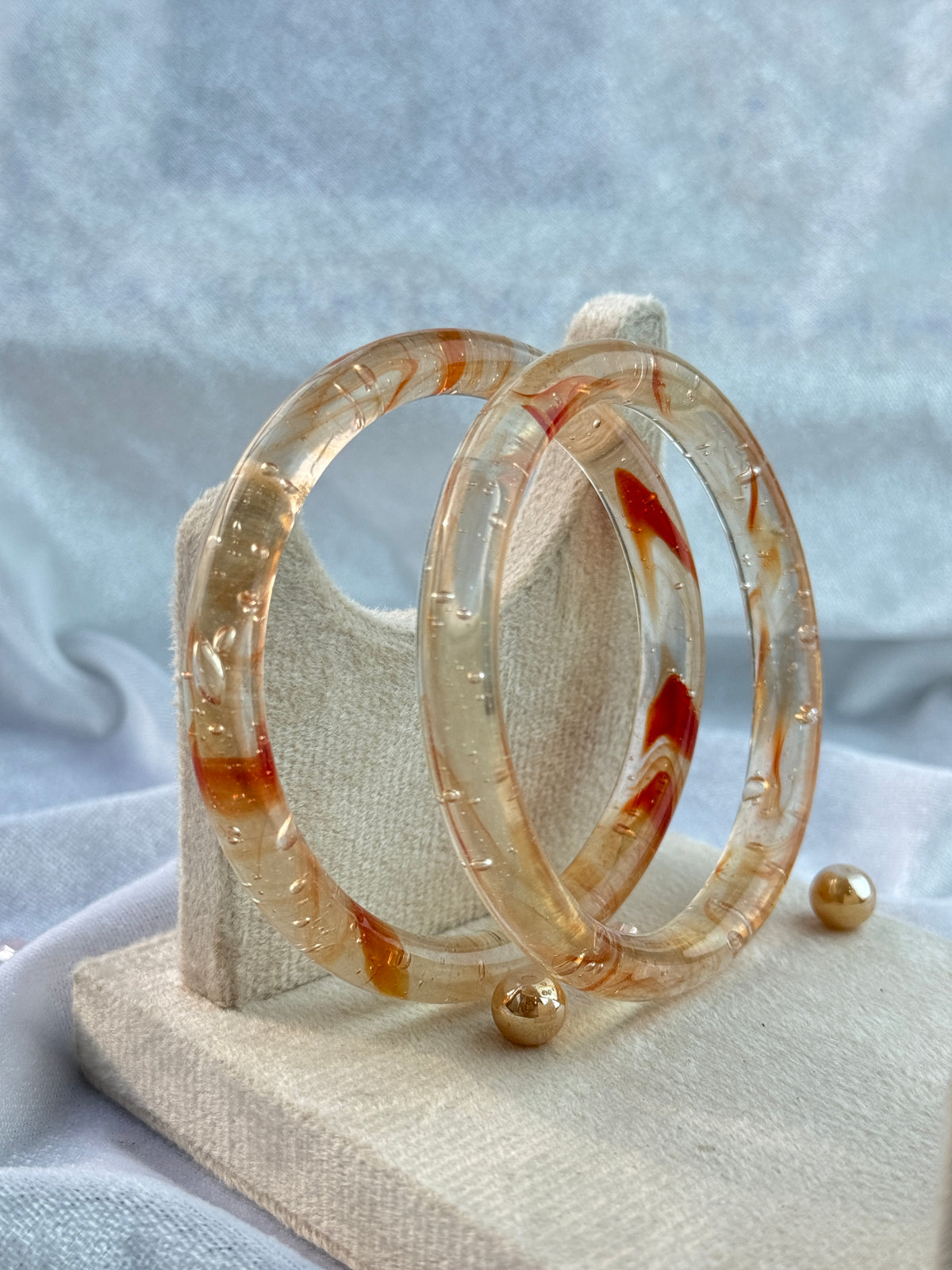 Two stacked translucent bangles with orange-brown marbled swirls, displayed on a textured cream fabric stand.