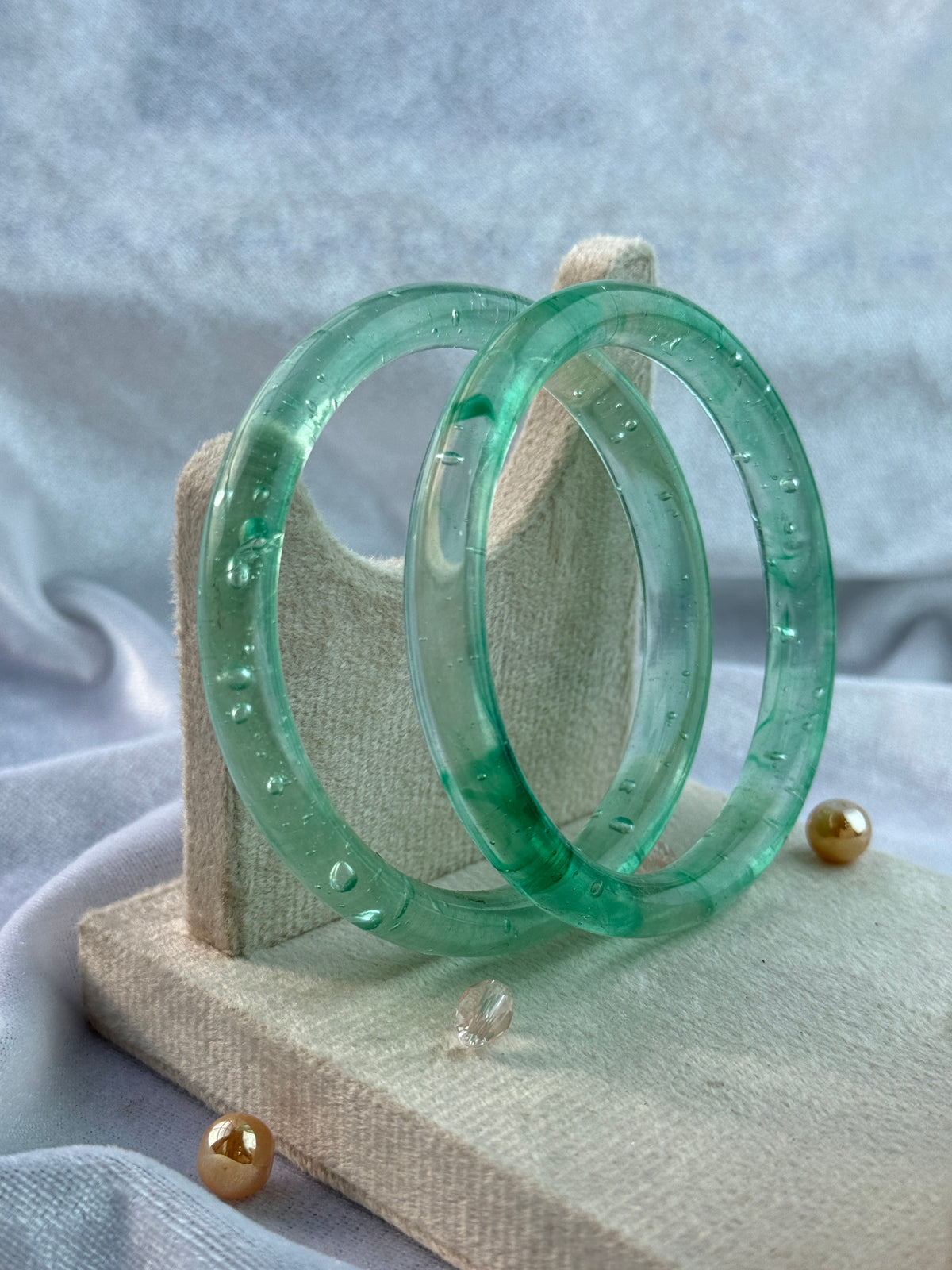 Two translucent light green glass bangles stacked on a beige fabric jewelry stand with gold-tone pins, set against a soft light blue-gray fabric background.