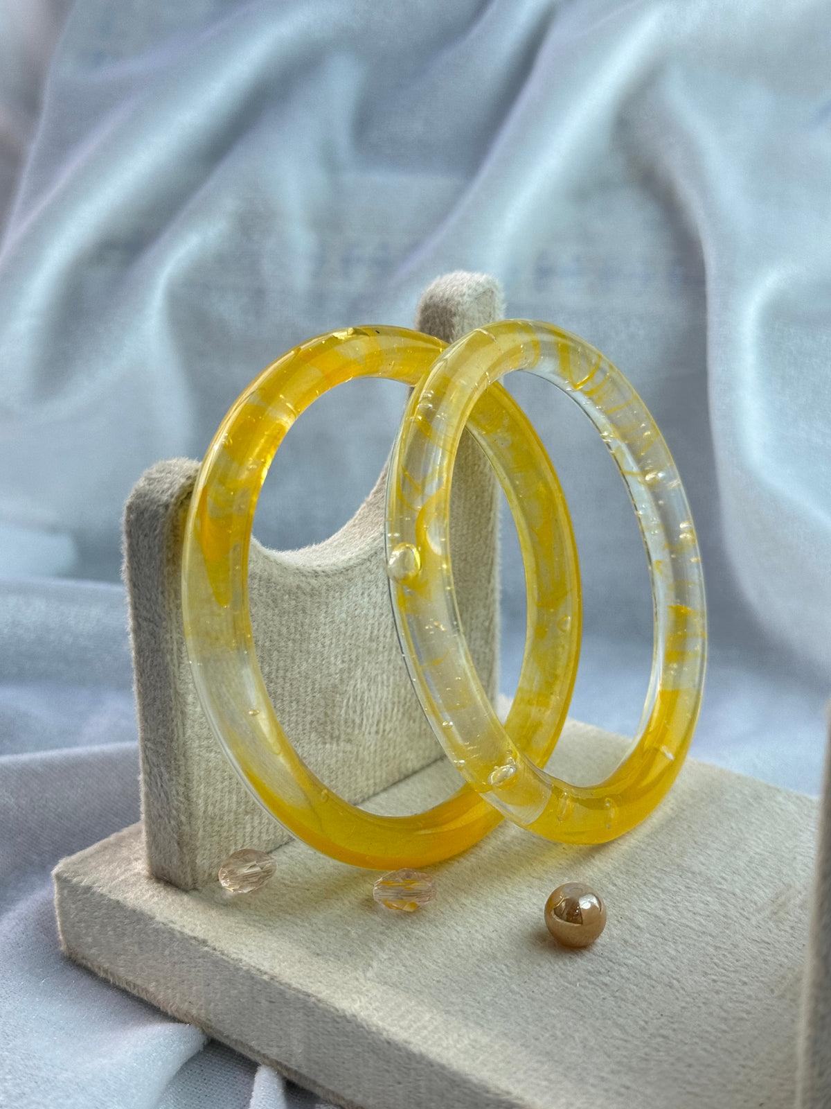Two translucent yellow resin bangles displayed on a small beige jewelry stand against a soft white fabric background.