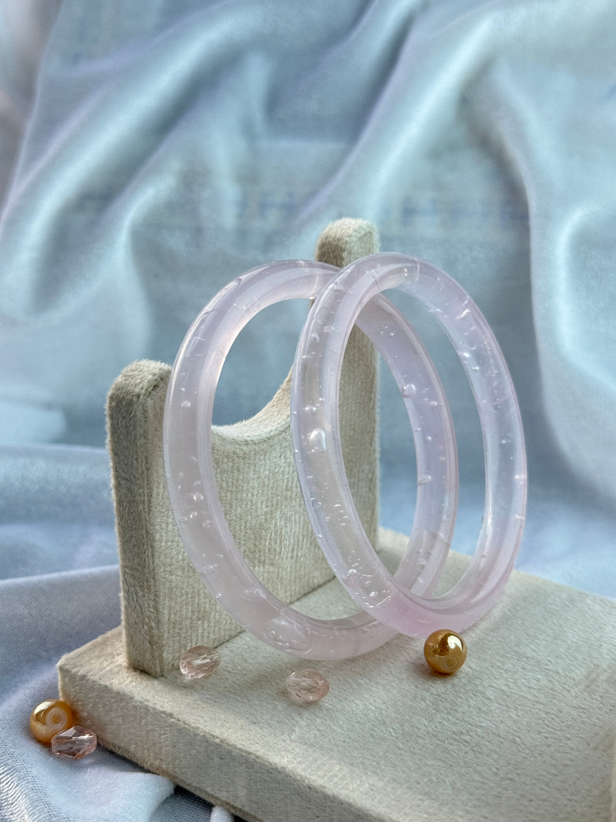 Two translucent pale pink bangles with tiny inlaid dots, displayed on a beige fabric stand against a soft light blue draped background.