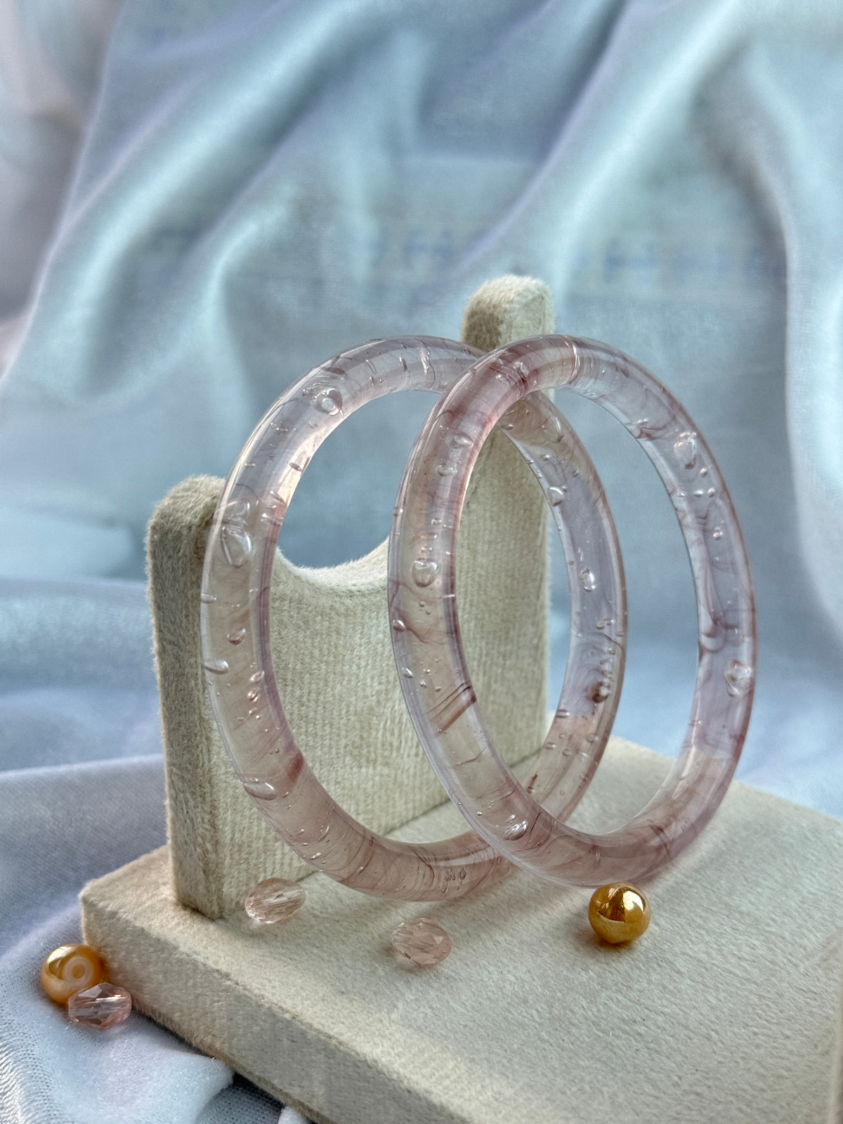Two translucent lavender-pink bangles with a textured, organic pattern, displayed on a beige fabric stand against a soft light blue satin background.