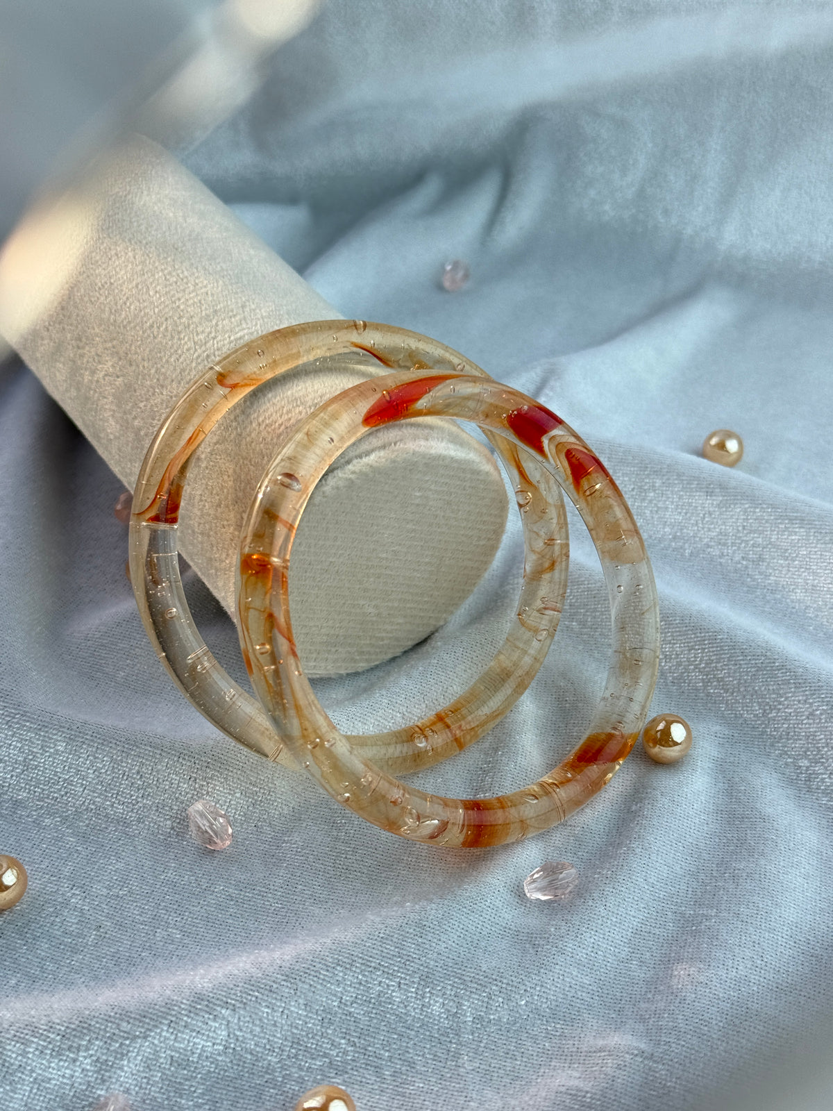 Three stacked translucent resin bangles with red and brown swirls, displayed on a white ring stand against a light blue satin fabric background.