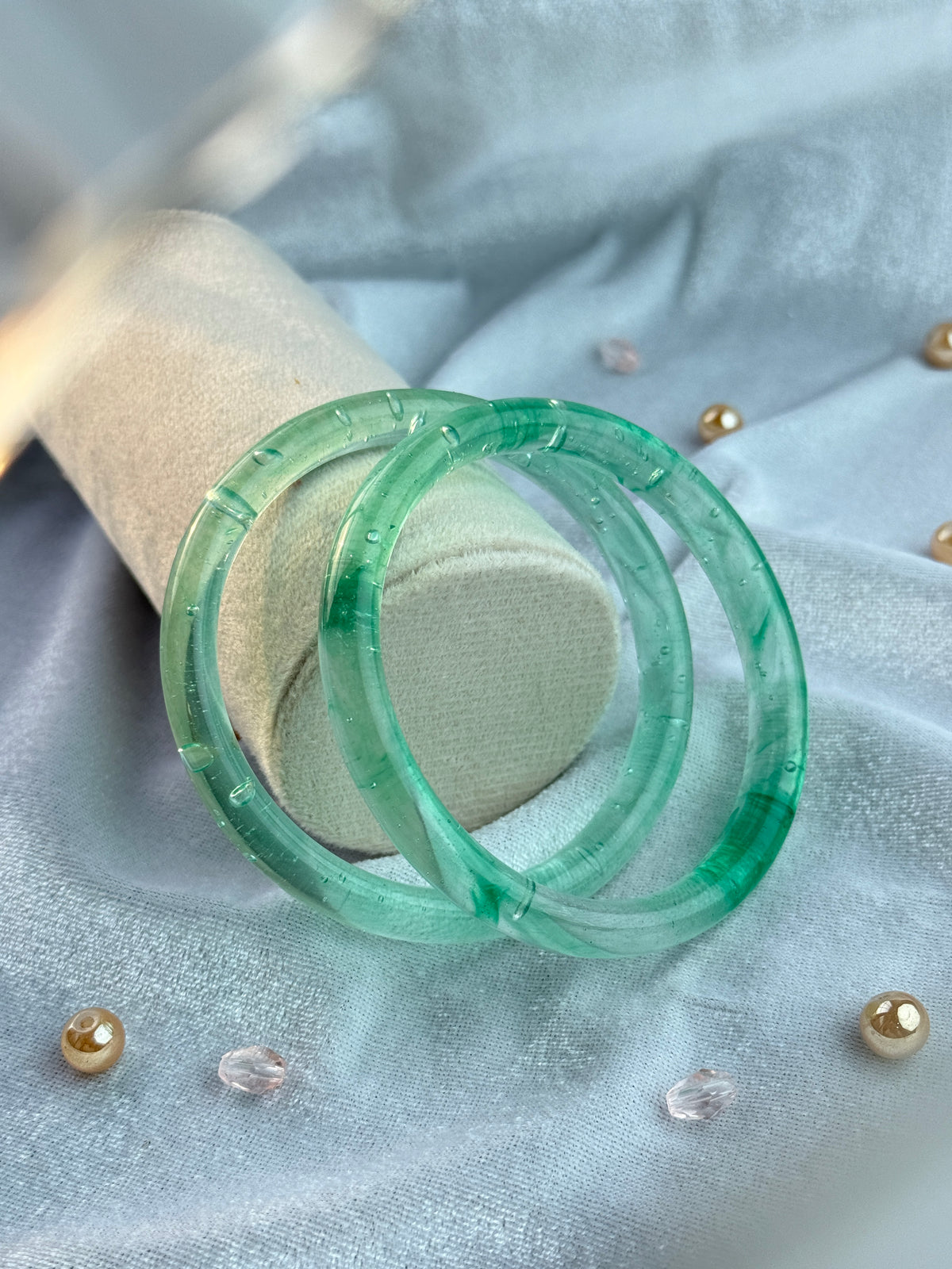Two translucent green bangles with a segmented design, displayed on a light beige fabric stand against a light blue satin background with gold and clear rhinestone embellishments.