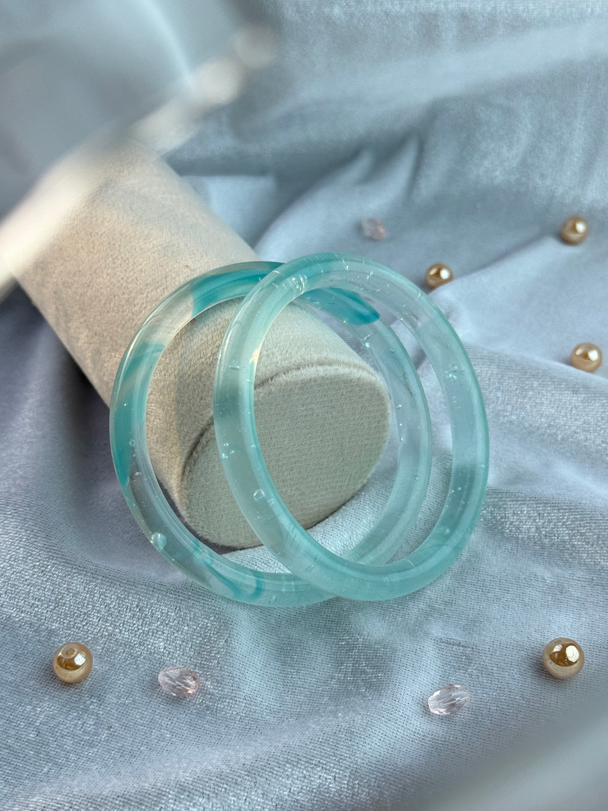 A translucent light blue-green acrylic ring displayed on a beige fabric ring stand, set against a light blue textured background with small gold and clear rhinestone embellishments.