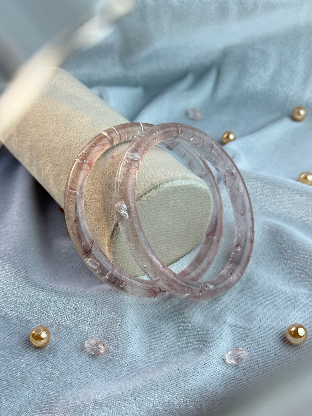 A pair of translucent light pink glass bangles displayed on a beige jewelry stand against a light blue fabric background with gold decorative accents.