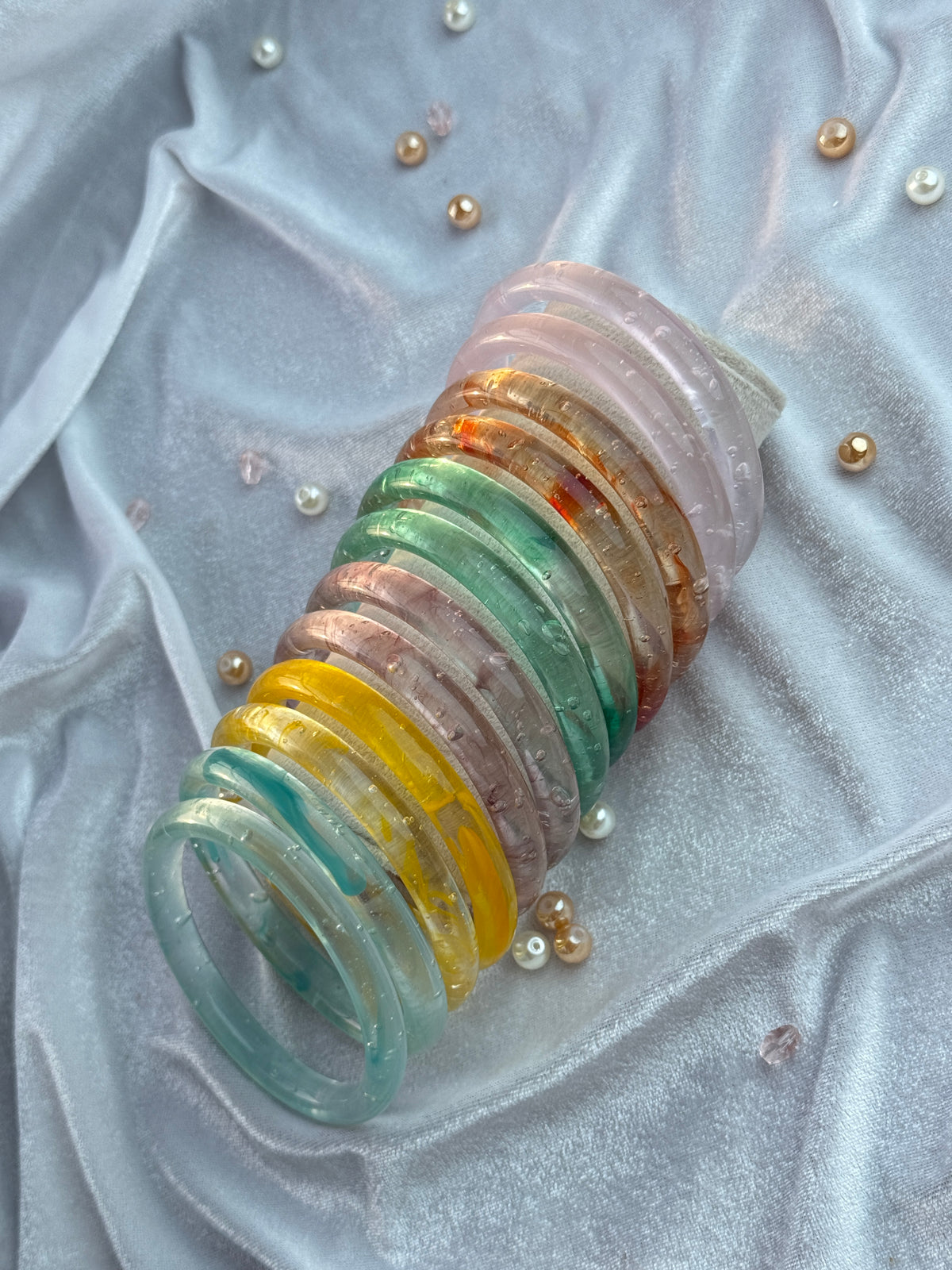 A stack of translucent glass bangles in assorted pastel and earthy colors, including pink, green, yellow, and brown, arranged on a draped light gray fabric with scattered decorative pearls.