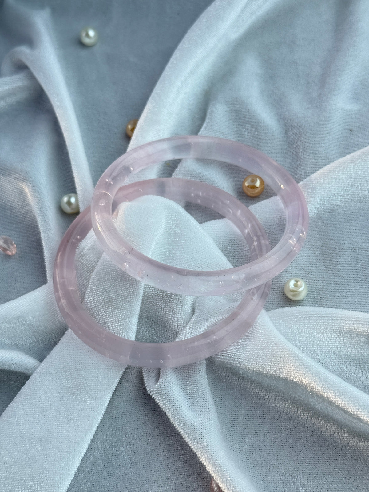 Two translucent light pink bangles with tiny inclusions, displayed on a draped light gray fabric background with small pearl and gold beads.