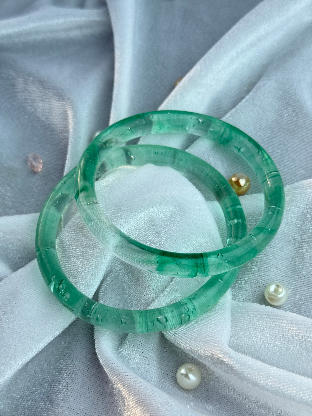 Two translucent green glass bangles with engraved characters, displayed on a light gray fabric background with pearl pins.