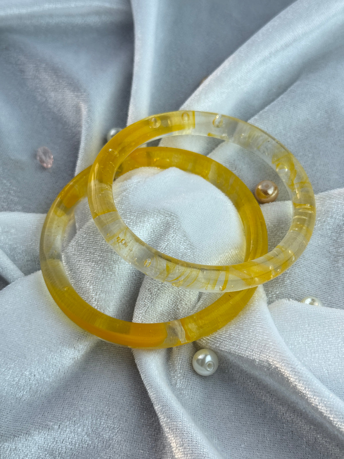 Two translucent yellow resin bangles with tiny inclusions, displayed on a light gray satin fabric background with pearl-like decorative pins.