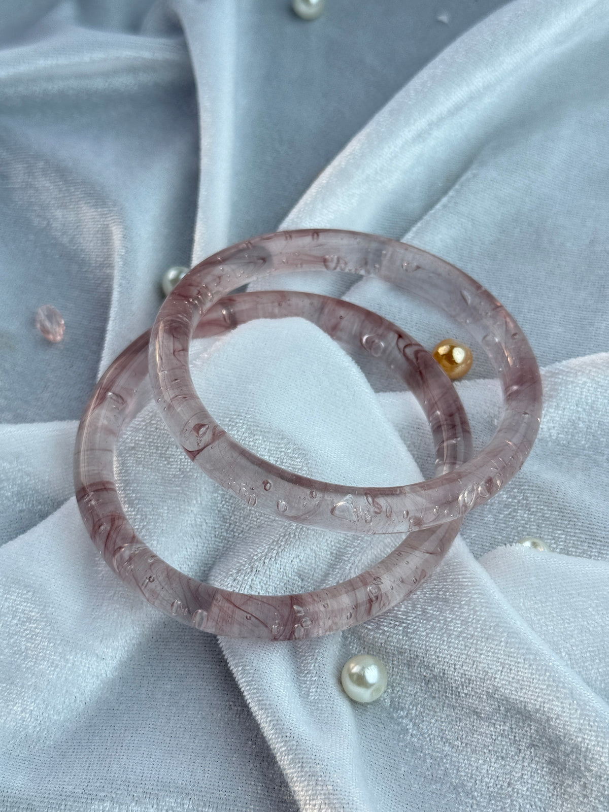 Two translucent pinkish-purple bangles with tiny clear inclusions and one small gold bead accent, displayed on draped light gray fabric with pearl-like beads.