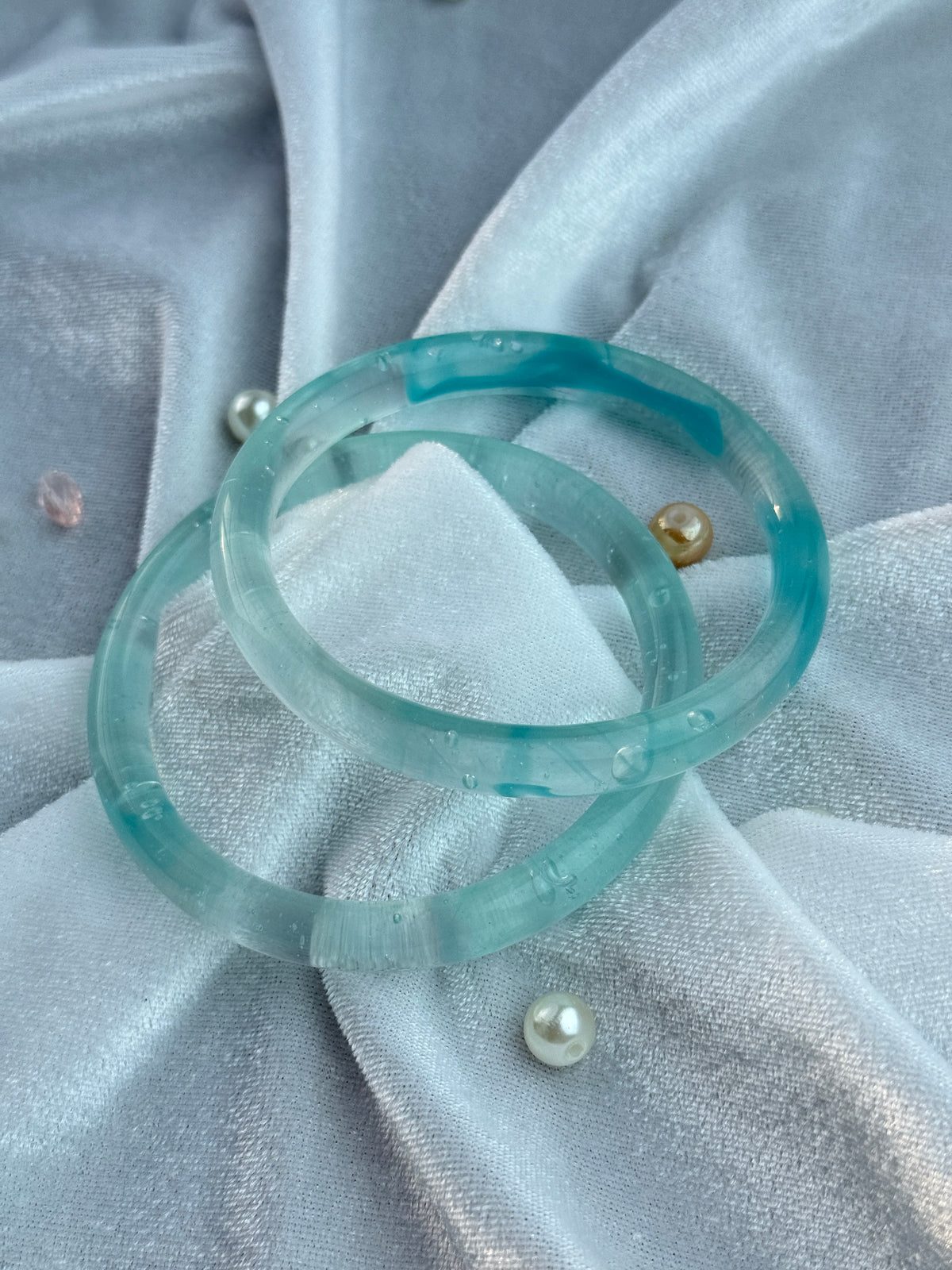 Two translucent aqua blue circular rings displayed on a light blue satin fabric background with small pearl-like beads.